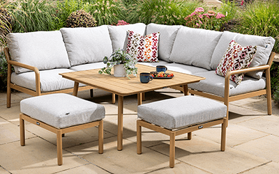 Image of Sahara square corner set with adjustable table. Light grey cushioned square sofa, and 2 stools, surround a height adjustable table, on an black and white print rug topped patio with countryside in background.