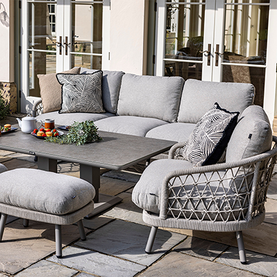 Image of Hartman Aries Lounge Set, with a 3 seat sofa, 2 armchairs and 2 stools, along with a rectangular table. Presented in a grey colourway, this set has rope weave seating areas, and thick cushions. Shown on a stone patio with house in background.,