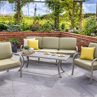 Image of Hartman Buxton Lounge Set, with a sofa, 2 chairs and rectangular table, in a patterned aluminium topped with green cushions, on a grey patio with a wall and garden behind.