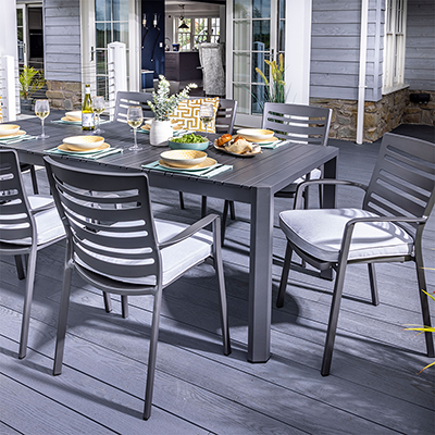 Image of Hartma 8 seat Dining Set, with dark grey slatted chair backs and table top, one chair pushed off at an angle to table. Set on grey patio with house at back.