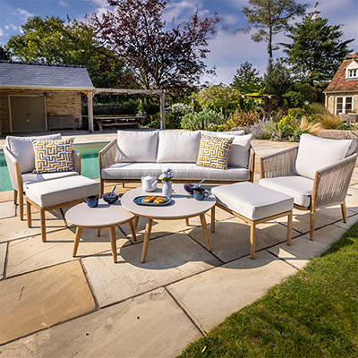 Image of Hartman Portico Lounge Set, with rope weave chair backs, light toned cushions on a faux teak frame, 2 round side tables, on a stone patio next to a pool in a garden