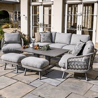 Image of Hartman Aries Lounge Set, with a 3 seat sofa, 2 armchairs and 2 stools, along with a rectangular table. Presented in a grey colourway, this set has rope weave seating areas, and thick cushions. Shown on a stone patio with house in background.,