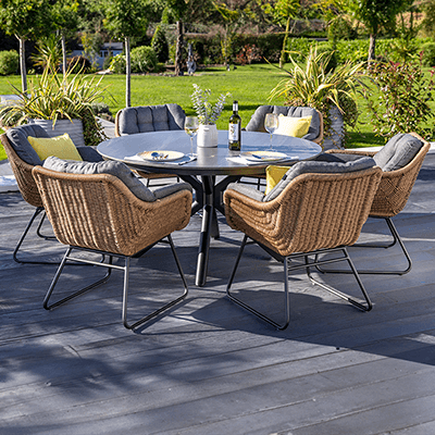 Image of Hartman Rayo Dining Set, featuring natural toned weave bucket chairs with cushions in a grey colourway, with a grey parasol, on a grey slat patio with countryside background