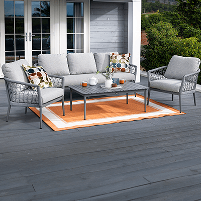 Image of Hartman Tierra Lounge Set, with geometric weave frame, featuring chair in foreground and sofa and part of table further back, in grey colourway, with cushions, on a grey slat patio with zebra print rug, and patio doors in background