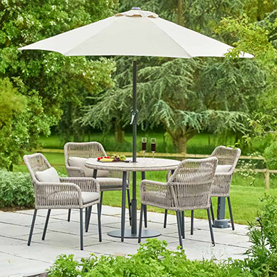Image of Panama 4 seat dining set with rope weave back chairs, with cream cushions, and a round table and parasol. Positioned on a stone patio, with trees in background