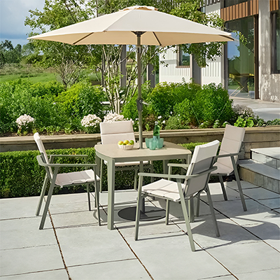 Image of LG Tuscany 4 seat dining set, with light green metal frame and cream seat pads, a round table and parasol, on a stone patio with a garden and house in the background