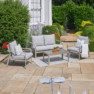 Image of LG Capri lounge set, with light grey metal frame and matching cushions, a rectangle coffee table, on a stone patio, with house and garden in background